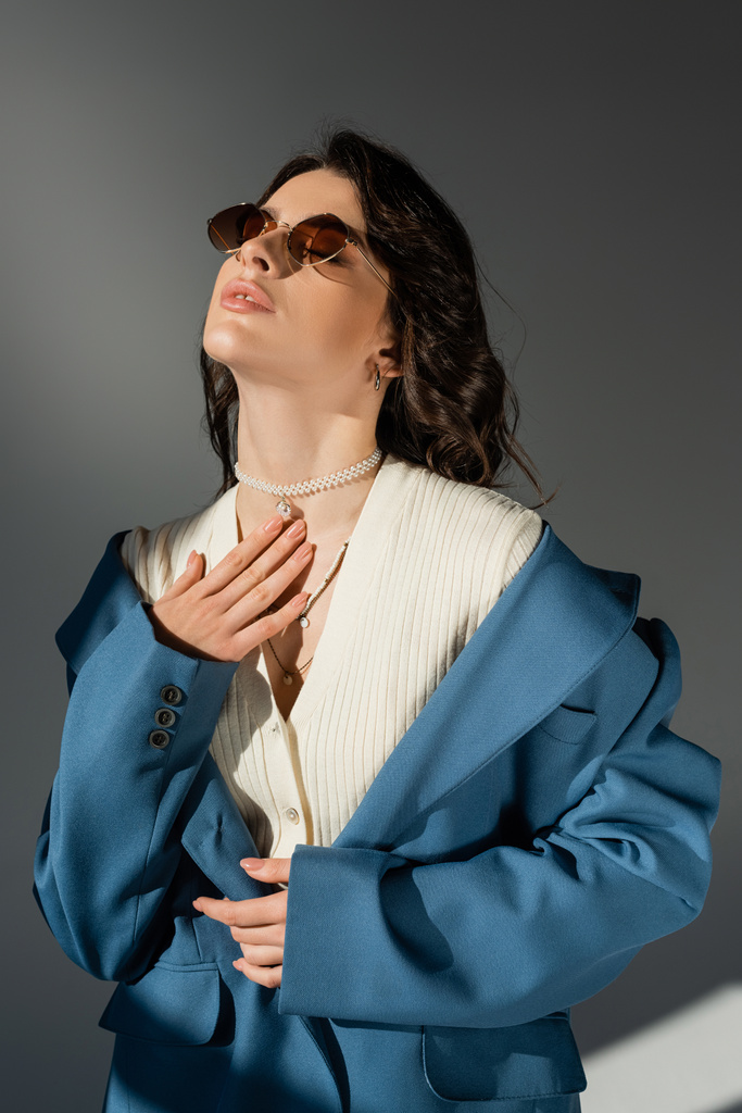 sinnliche Frau in pastellblauer Jacke und stylischer Sonnenbrille posiert mit der Hand auf der Brust auf grauem Hintergrund - Foto, Bild