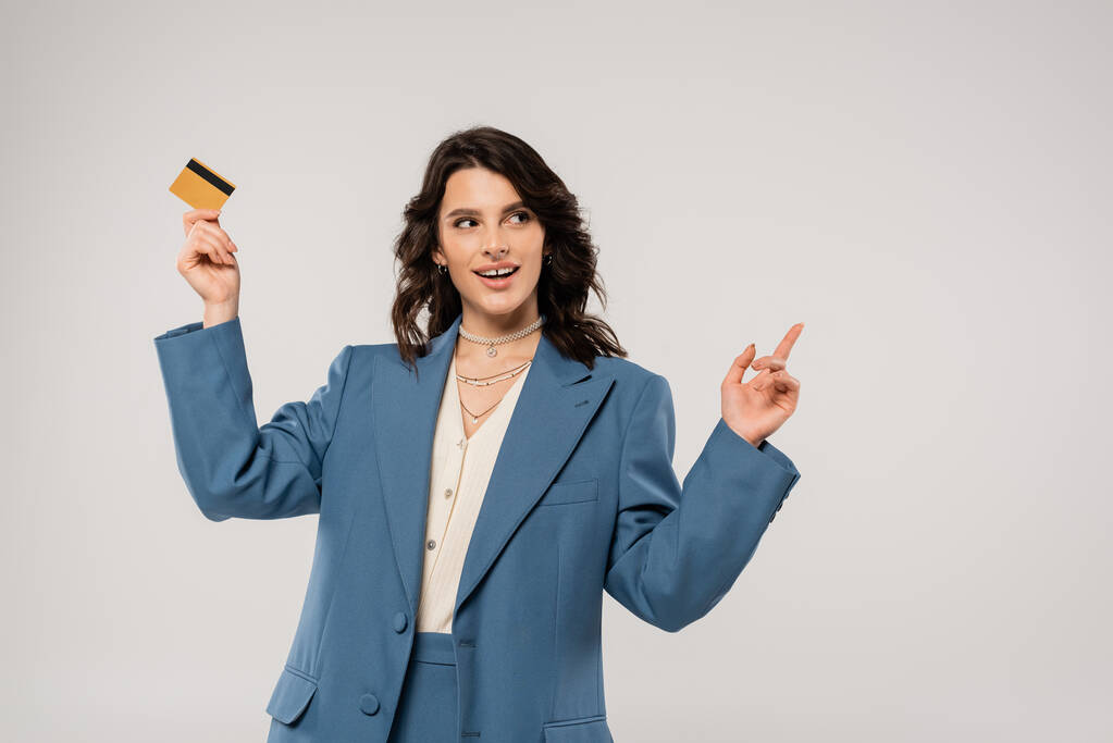 задоволена жінка в блакитному блістері тримає кредитну картку і вказує пальцем ізольовано на сірому
 - Фото, зображення