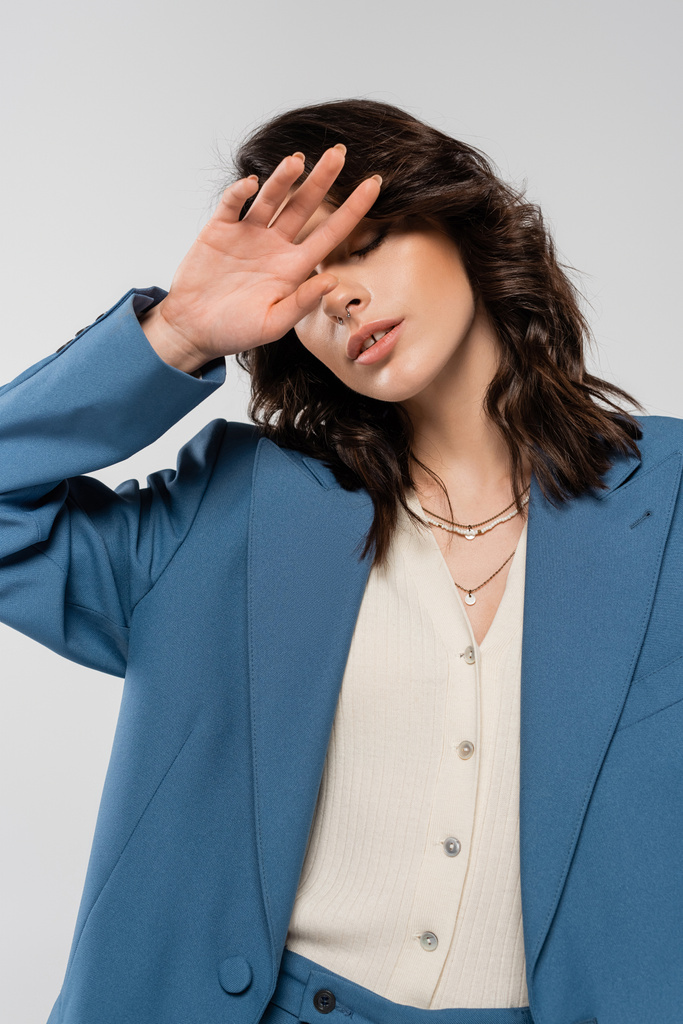 divatos nő kék dzsekiben és nyakláncokkal borító arc kézzel elszigetelt szürke - Fotó, kép
