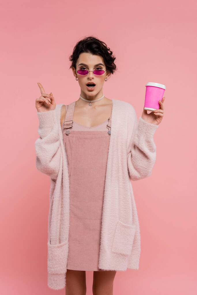 femme étonnée dans un cardigan chaud et des lunettes de soleil tenant tasse en papier et pointant avec le doigt isolé sur rose - Photo, image