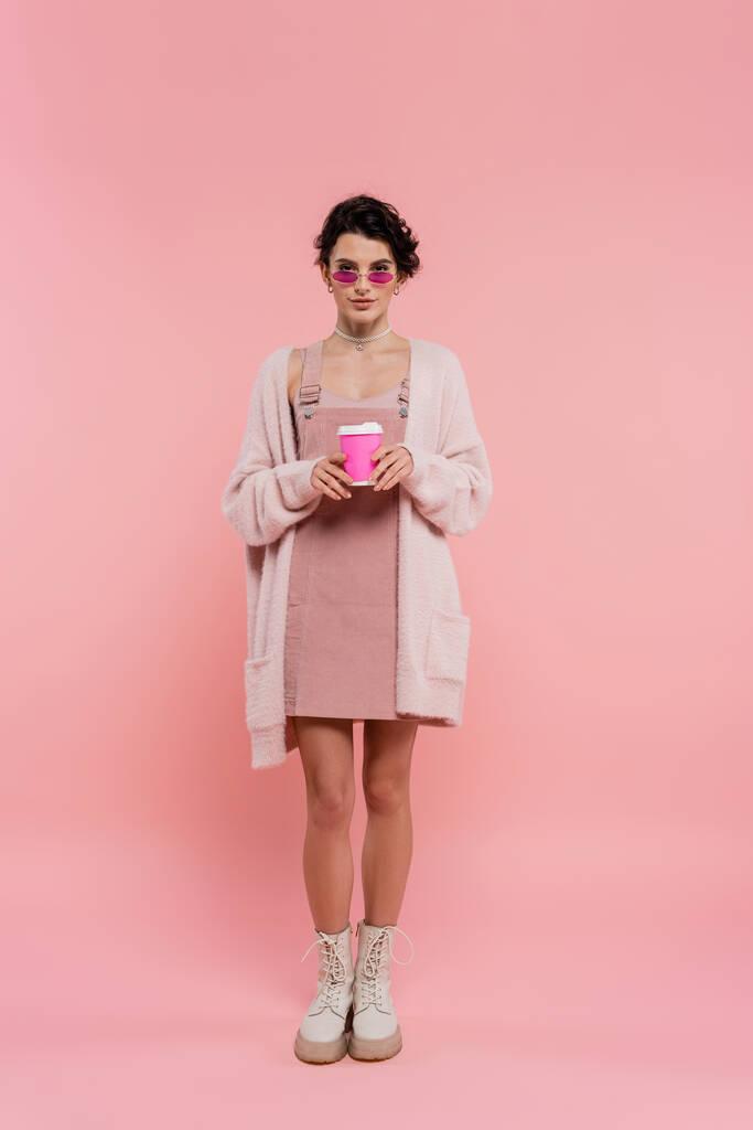 pleine longueur de femme en cardigan chaud et des lunettes de soleil à la mode tenant du café pour aller sur rose  - Photo, image
