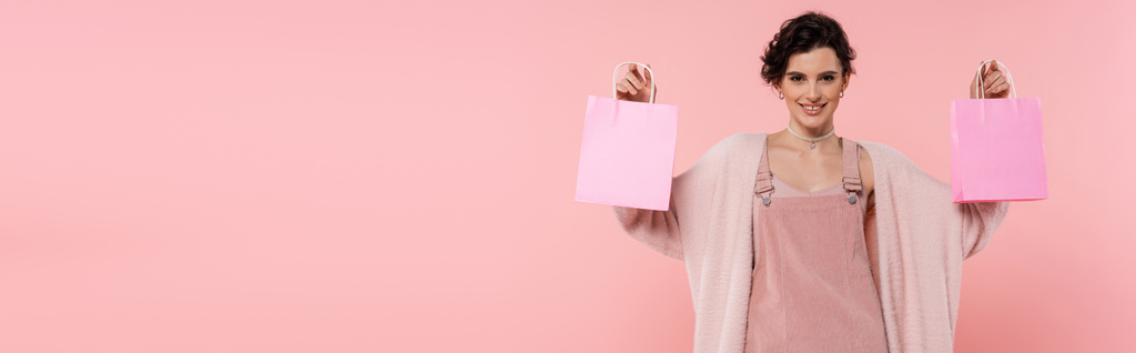 boldog barna nő meleg kardigán mutatja bevásárló táskák elszigetelt rózsaszín, banner - Fotó, kép