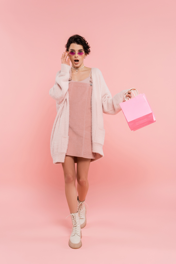 μοντέρνα και έκπληκτος γυναίκα αγγίζοντας γυαλιά ηλίου, ενώ το περπάτημα με τσάντες ψώνια σε ροζ  - Φωτογραφία, εικόνα