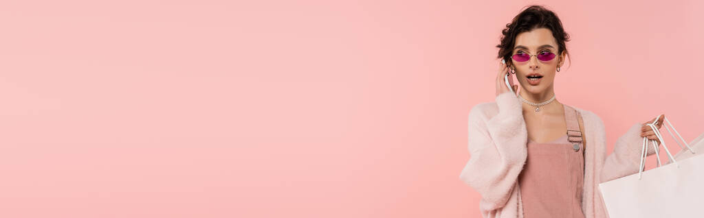donna bruna in cardigan caldo e occhiali da sole che tiene le borse della spesa e parla su smartphone isolato su rosa, banner - Foto, immagini
