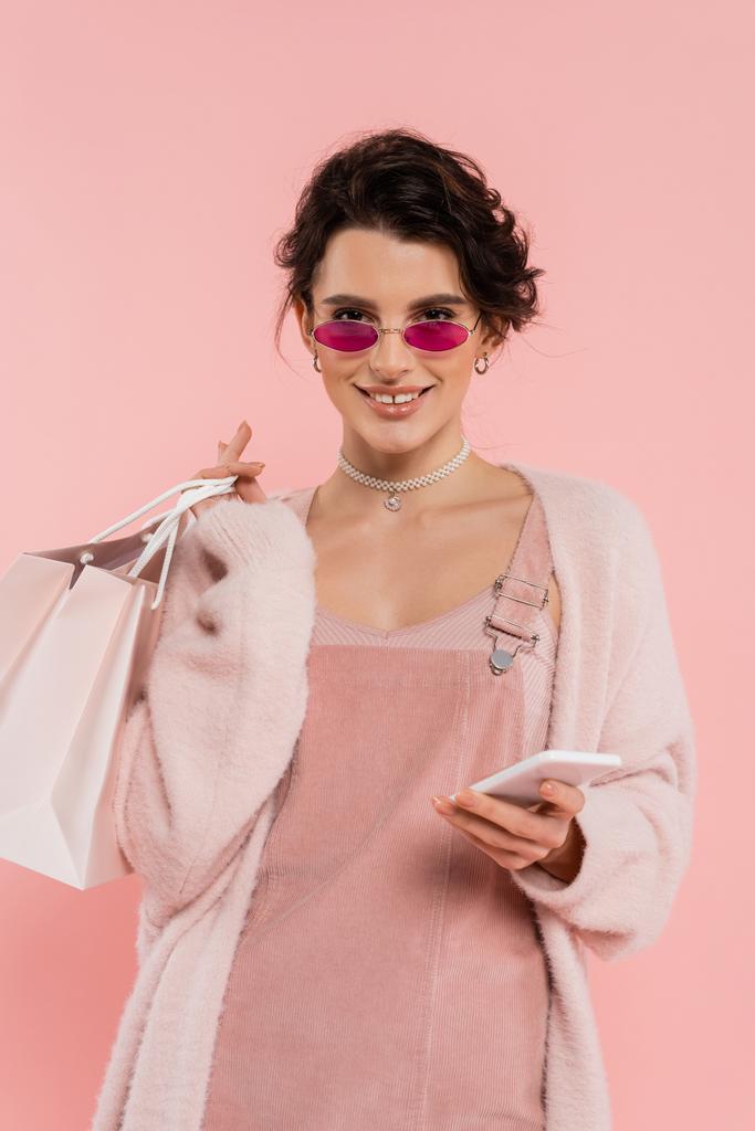 mulher feliz e elegante com sacos de compras e telefone celular olhando para a câmera isolada em rosa - Foto, Imagem