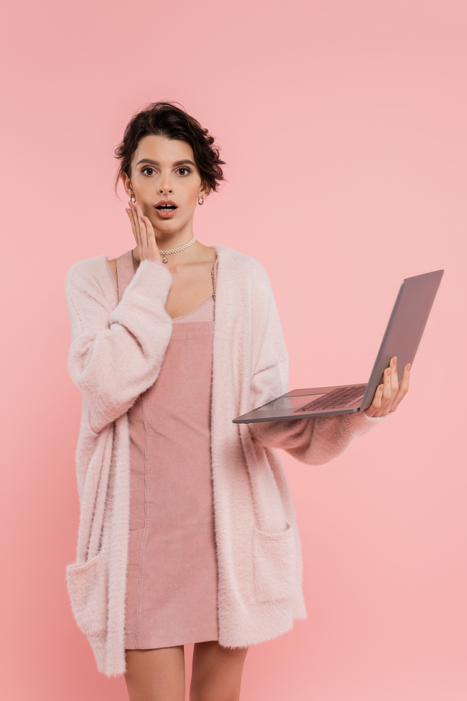 Besorgte Frau mit Laptop berührt Gesicht und blickt in Kamera isoliert auf rosa - Foto, Bild