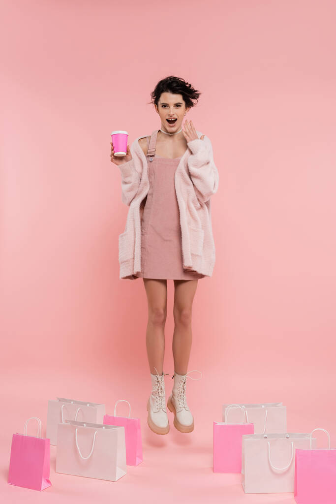 comprimento total da mulher surpreendida com café para ir levitar perto de sacos de compras no fundo rosa - Foto, Imagem