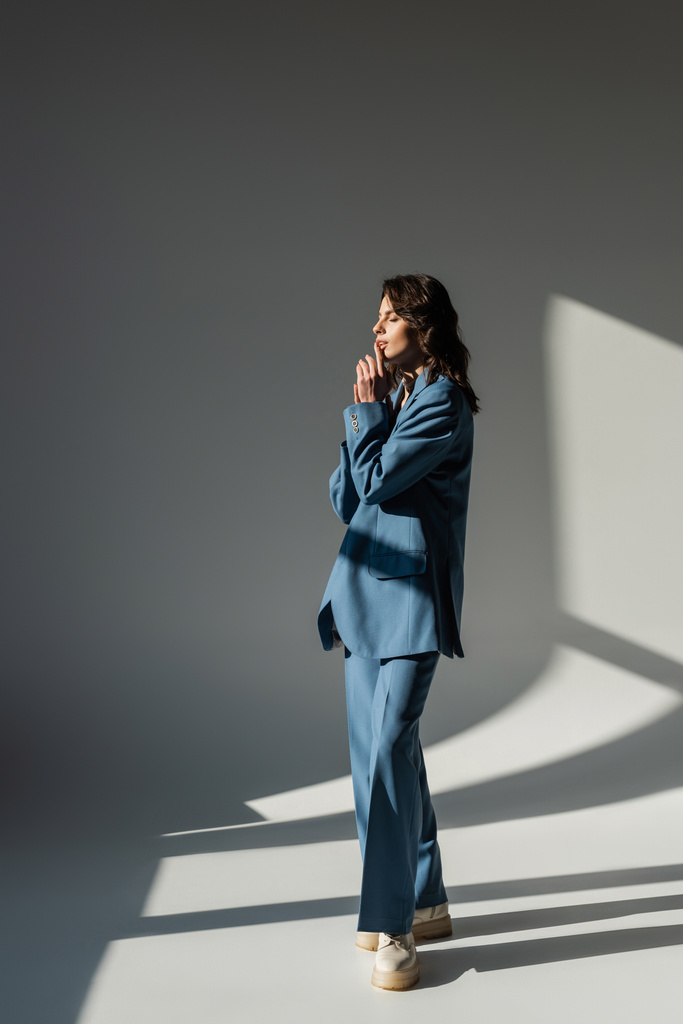полная длина модной женщины в синем костюме стоящей с закрытыми глазами на сером фоне с освещением - Фото, изображение