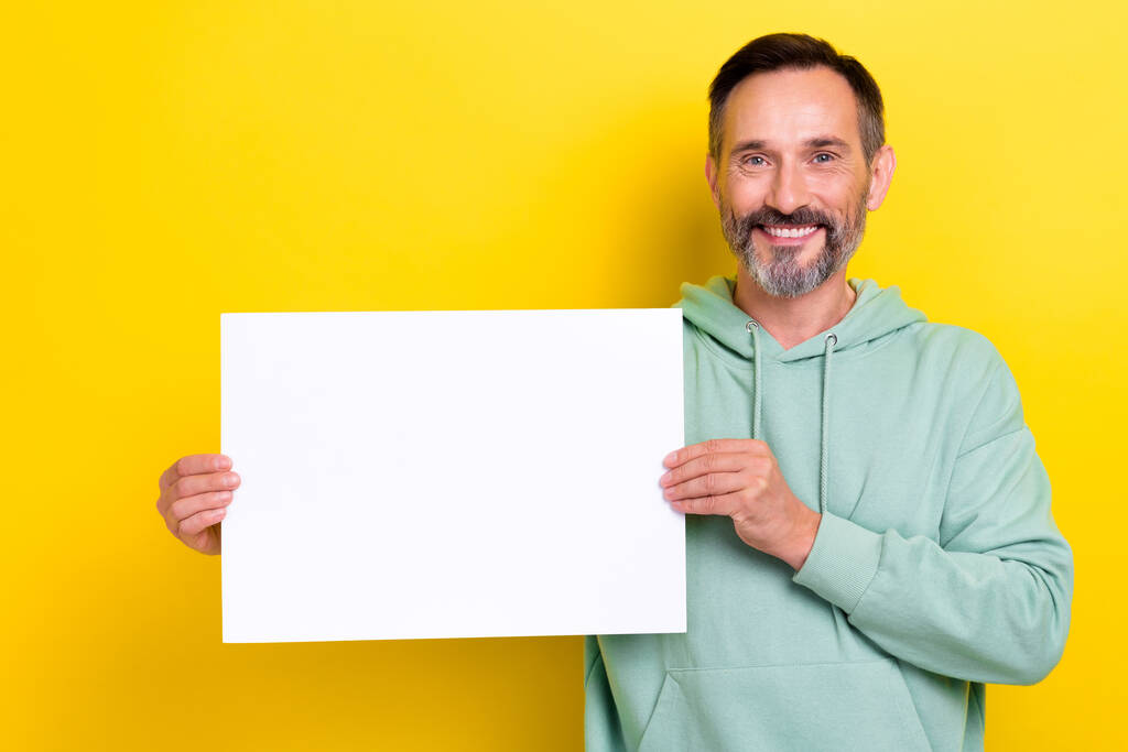 Foto van gepensioneerde middelbare leeftijd brunet haar vrolijke glimlachende man houden papier poster lege ruimte advertentie nieuw merk geïsoleerd op gele kleur achtergrond. - Foto, afbeelding
