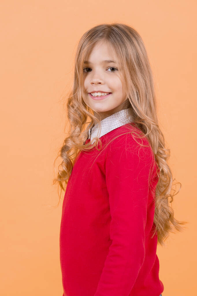 Child with curly blond hair in red sweater. Girl smile on orange background. Happy childhood concept. Beauty, look, hairstyle. Kid fashion and style. - Photo, Image