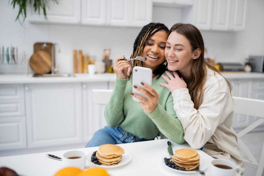 allegra donna lesbica africana americana mostrando smartphone alla fidanzata felice durante la colazione  - Foto, immagini