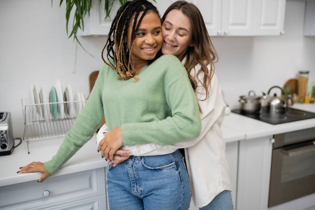 Šťastné lesbičky žena objímající radostné africké americké přítelkyně v kuchyni  - Fotografie, Obrázek