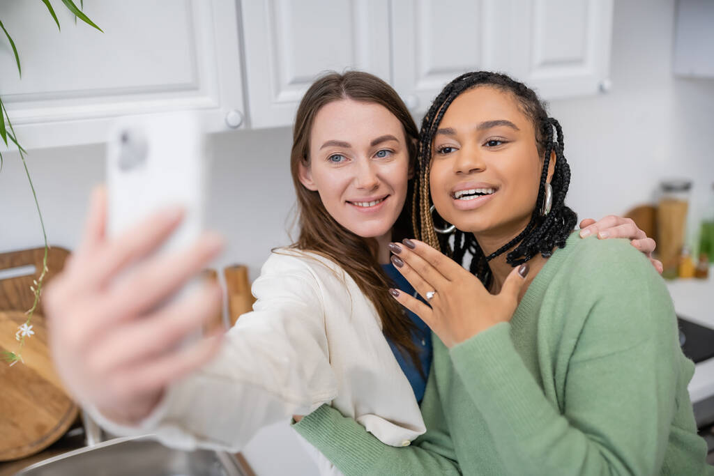 lesbische Frau macht Selfie, während fröhliche afrikanisch-amerikanische Freundin Verlobungsring am Finger zeigt  - Foto, Bild