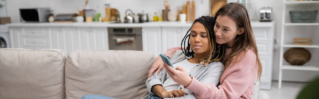 νεαρή λεσβία γυναίκα μηνυμάτων στο smartphone, ενώ στηρίζεται στον καναπέ με την αφρικανική αμερικανική φίλη, πανό  - Φωτογραφία, εικόνα