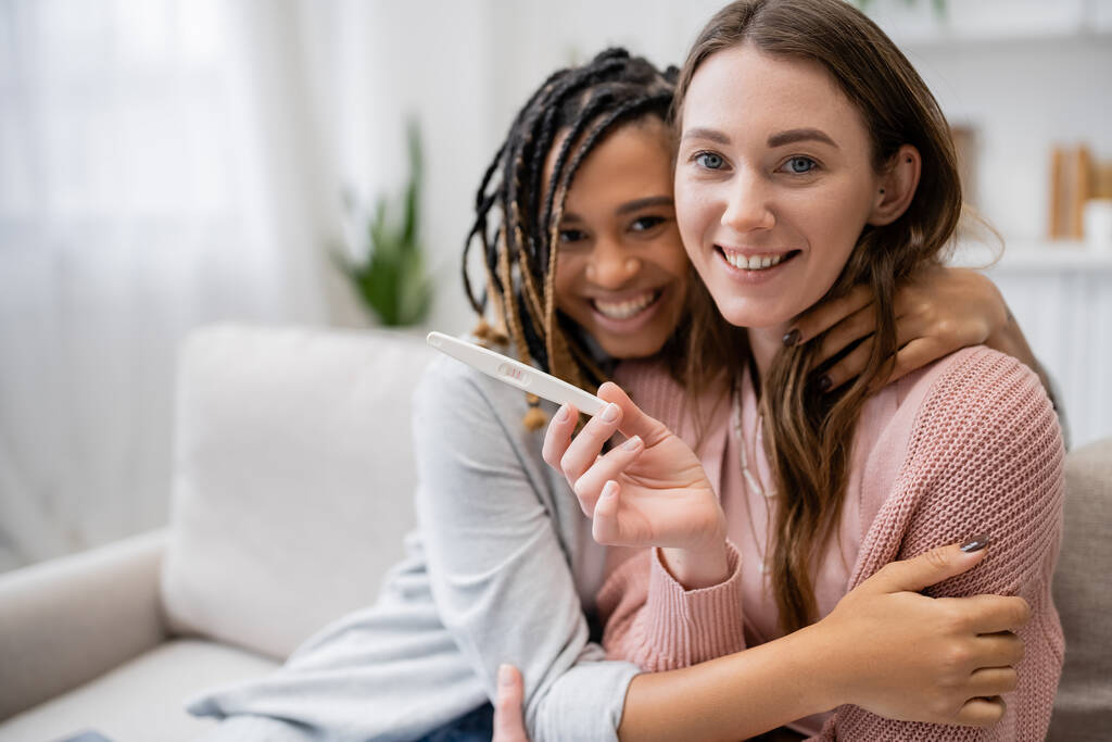 cheerful african american and lesbian woman hugging happy girlfriend with pregnancy test - Photo, Image