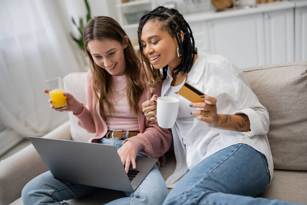 жизнерадостная американская лесбиянка из Африки держит кредитку рядом с счастливой девушкой, пользуясь ноутбуком во время покупок в Интернете  - Фото, изображение