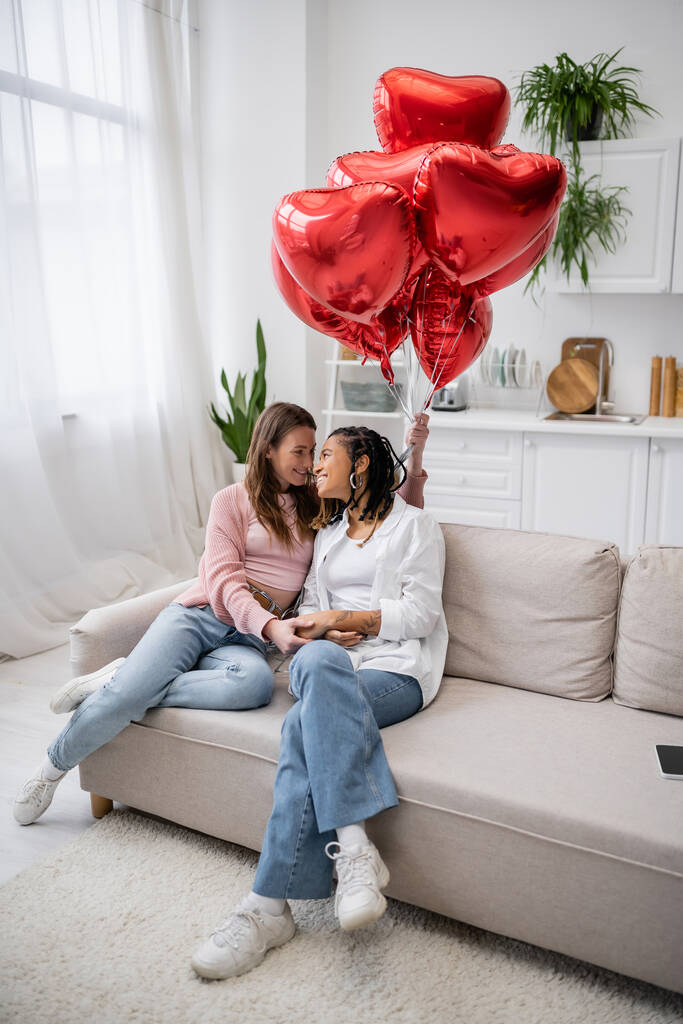 joyeuse femme lesbienne tenant des ballons en forme de coeur et assise sur le canapé avec une petite amie afro-américaine  - Photo, image