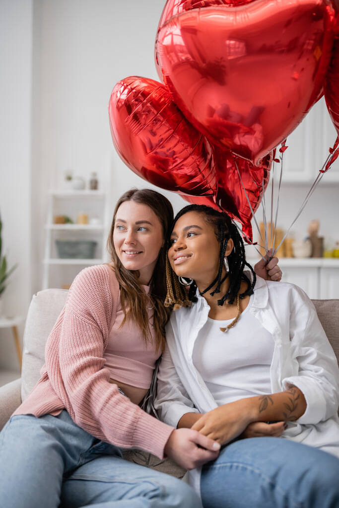 šťastná lesbička drží srdce ve tvaru balónky a sedí na gauči s tetovanou africkou americkou přítelkyní - Fotografie, Obrázek