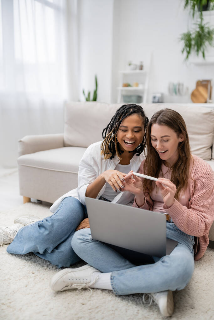 happy and lesbian woman showing pregnancy test near african american girlfriend during video call on laptop - Photo, Image