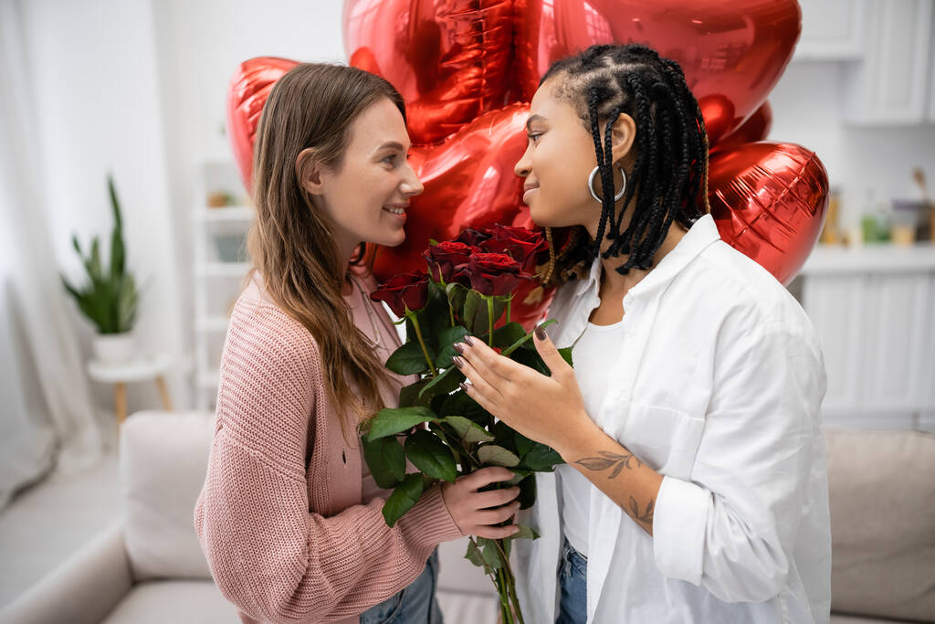 улыбающаяся лесбиянка с красными розами и смотрящая на татуированную африканскую подружку рядом с воздушными шарами  - Фото, изображение