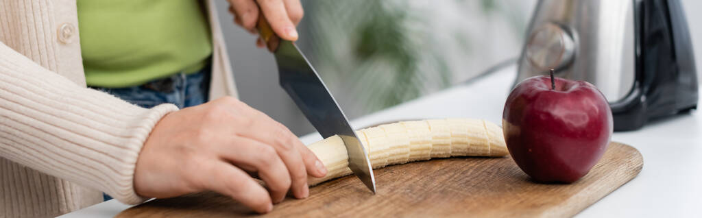 Vista recortada de la mujer cortando plátano fresco cerca de manzana y licuadora en la cocina, pancarta  - Foto, imagen