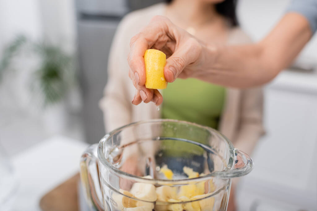 Ausgeschnittene Ansicht einer Frau, die in der Küche Zitrone über Mixer mit Früchten auspresst  - Foto, Bild