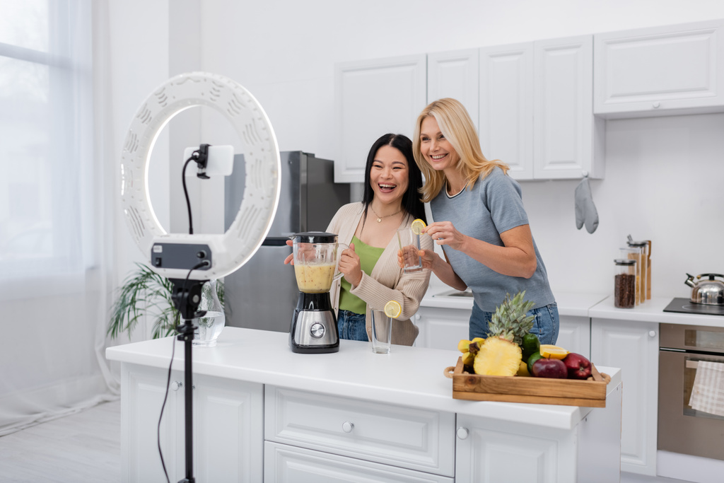 Fröhliche multiethnische Frauen halten Smoothie und Glas neben Smartphone mit Ringlampe in der Küche  - Foto, Bild