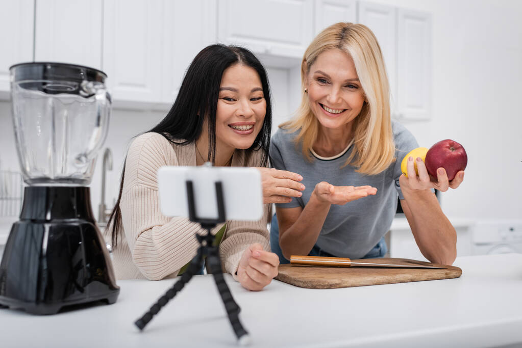 Mulheres inter-raciais positivas apontando para frutas perto do liquidificador e smartphone no tripé na cozinha  - Foto, Imagem