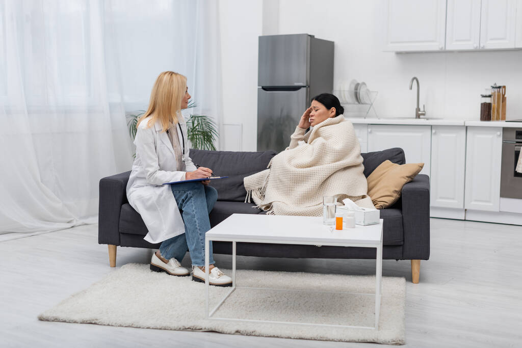 Kranke asiatische Frau in Decke sitzt in der Nähe von Arzt mit Klemmbrett zu Hause  - Foto, Bild