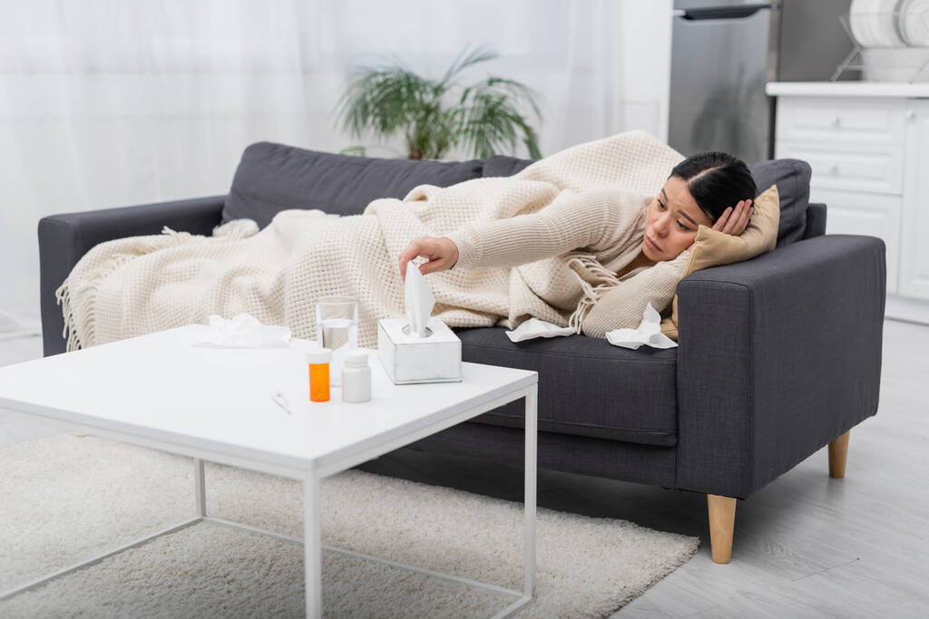 Άρρωστη Ασιάτισσα που παίρνει χαρτοπετσέτα κοντά σε χάπια και θερμόμετρο στο σπίτι  - Φωτογραφία, εικόνα