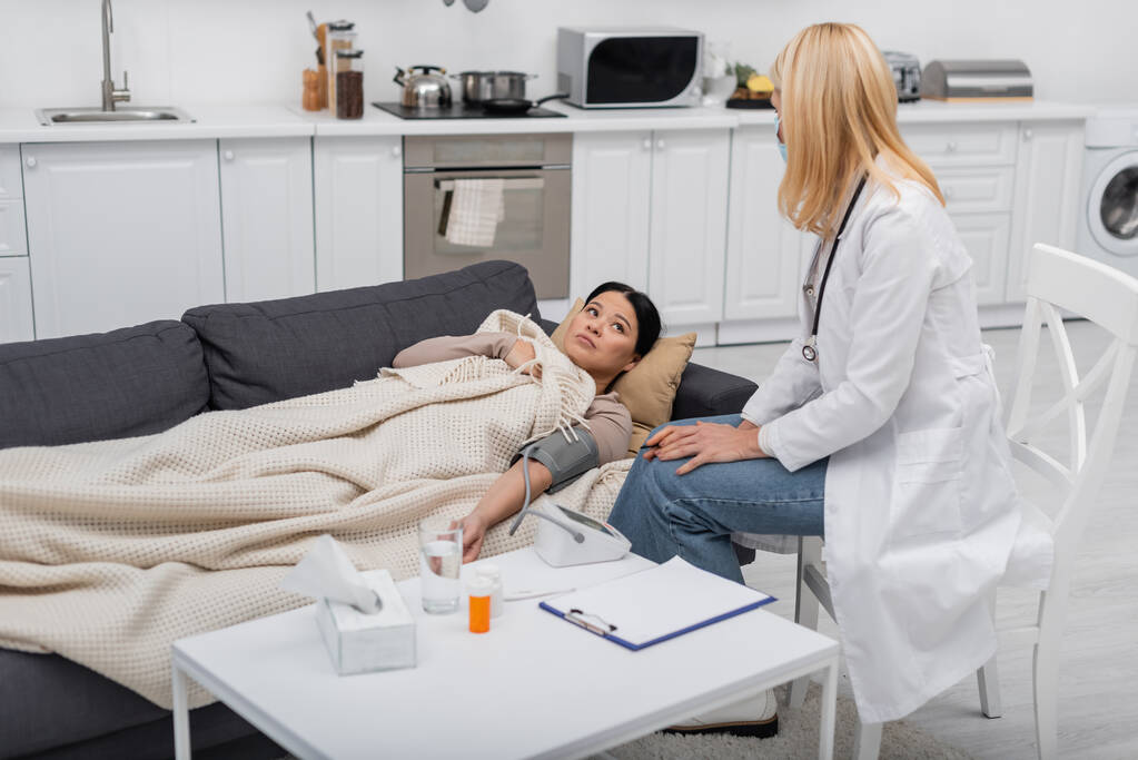 Malade asiatique patient couché près du médecin dans le masque médical et tonomètre à la maison  - Photo, image