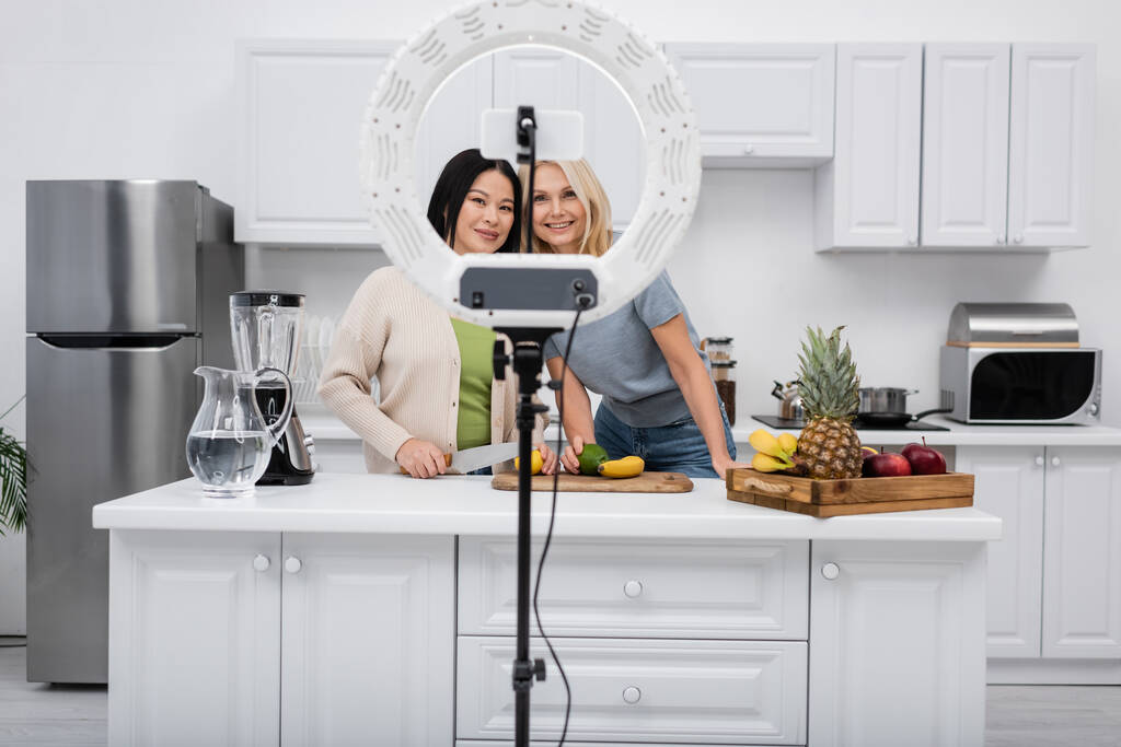 Bloggers interracial positivos mirando el smartphone en la lámpara de anillo cerca de frutas en la cocina  - Foto, Imagen