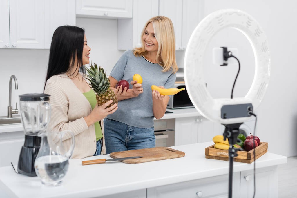 Międzyrasowi blogerzy uśmiechający się i trzymający owoce w pobliżu smartfona w lampie pierścieniowej w kuchni  - Zdjęcie, obraz