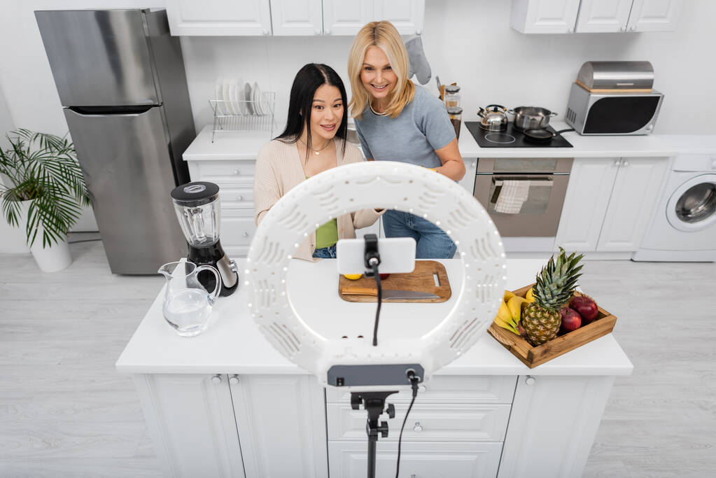 Межрасовые друзья смотрят на смартфон в кольцевой лампе возле блендера и фрукты на кухне  - Фото, изображение