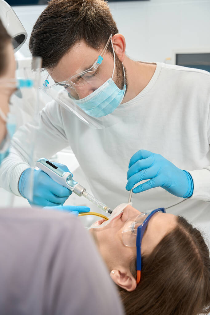 Lääkäri kertakäyttöisissä nitriilikäsineissä ruiskuttamassa anestesia-ainetta asiakkaalle hammasruiskupistoolilla - Valokuva, kuva