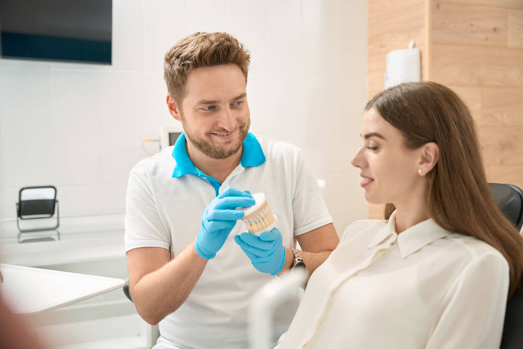 Hymyilevä ystävällinen hammaslääkäri osoittaa typodont nuorelle naispotilaalle, joka istuu hammastuolissa - Valokuva, kuva