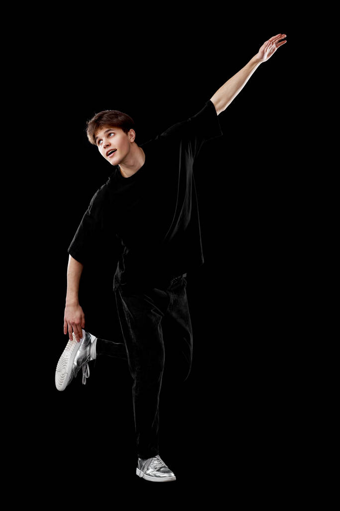 Nuori mies mustassa t-paidassa tanssii hassusti tehden liikkeitä mustalla pohjalla. Täyspitkä - Valokuva, kuva