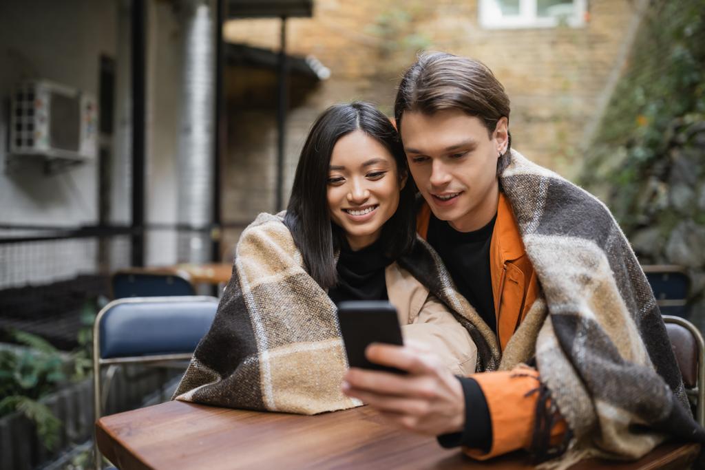 Sonriente pareja interracial en manta usando smartphone en cafetería al aire libre  - Foto, imagen