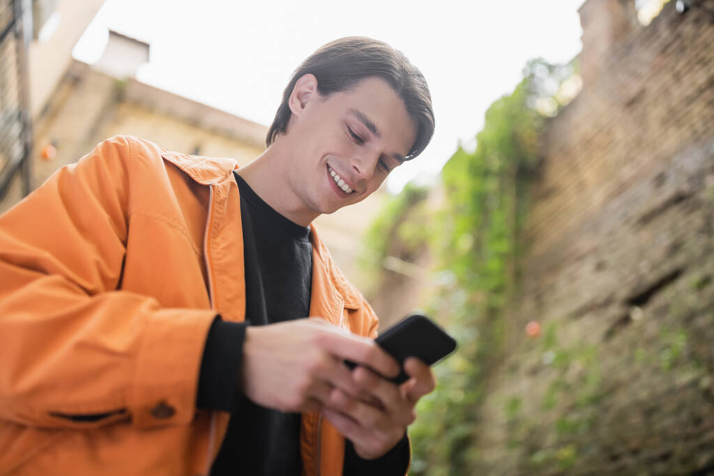Visão de baixo ângulo do homem sorridente na jaqueta usando o telefone móvel ao ar livre  - Foto, Imagem