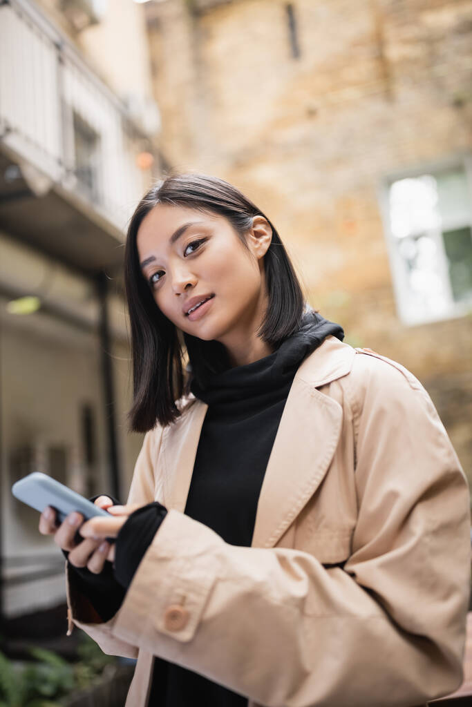 Femme asiatique élégante en trench coat tenant smartphone et regardant la caméra à l'extérieur  - Photo, image