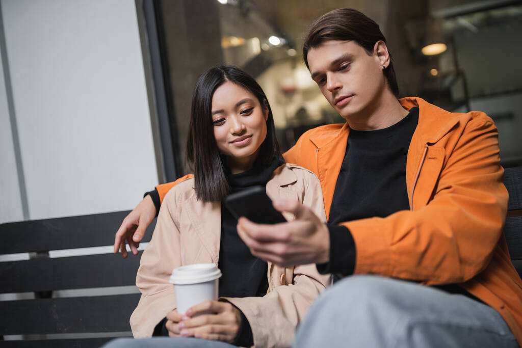 jong multi-etnische paar met koffie te gaan en smartphone zitten op de bank op het terras van cafe  - Foto, afbeelding
