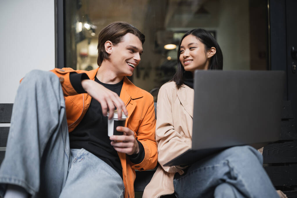 Couple multiethnique souriant avec café à emporter et ordinateur portable se regardant mutuellement près du café  - Photo, image