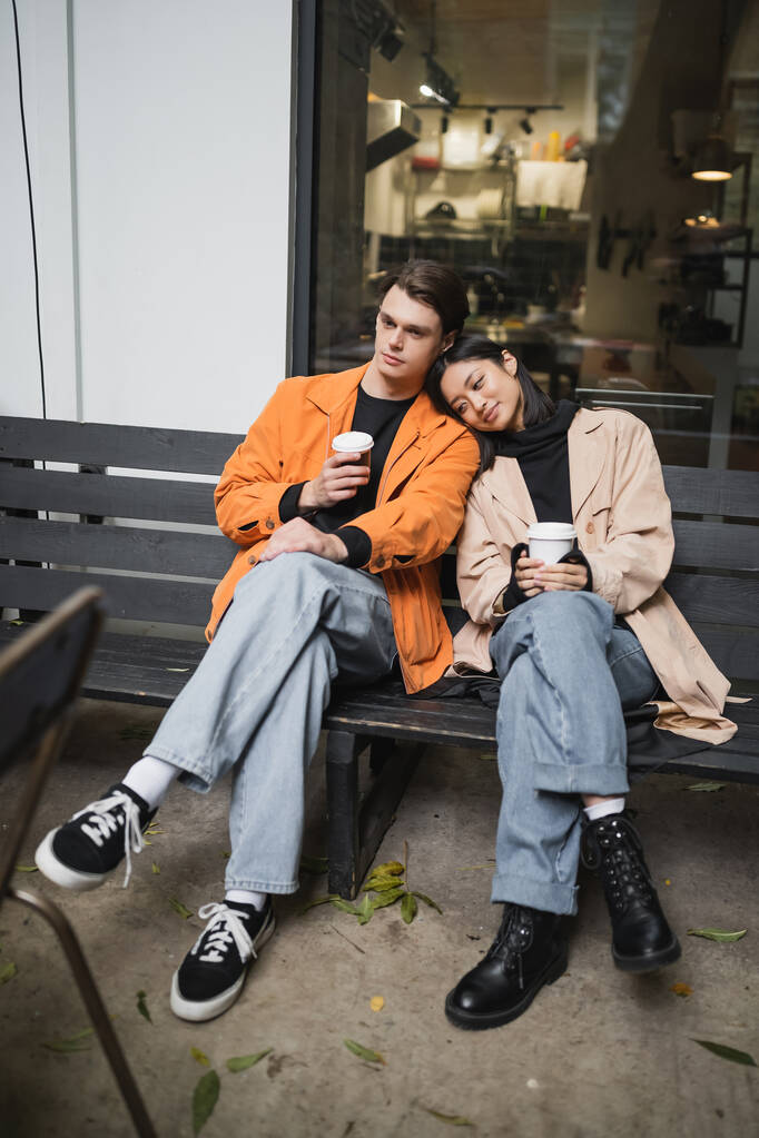 Junge Asiatin im Trenchcoat hält Kaffee in der Nähe ihres Freundes auf Bank in der Nähe des Cafés  - Foto, Bild