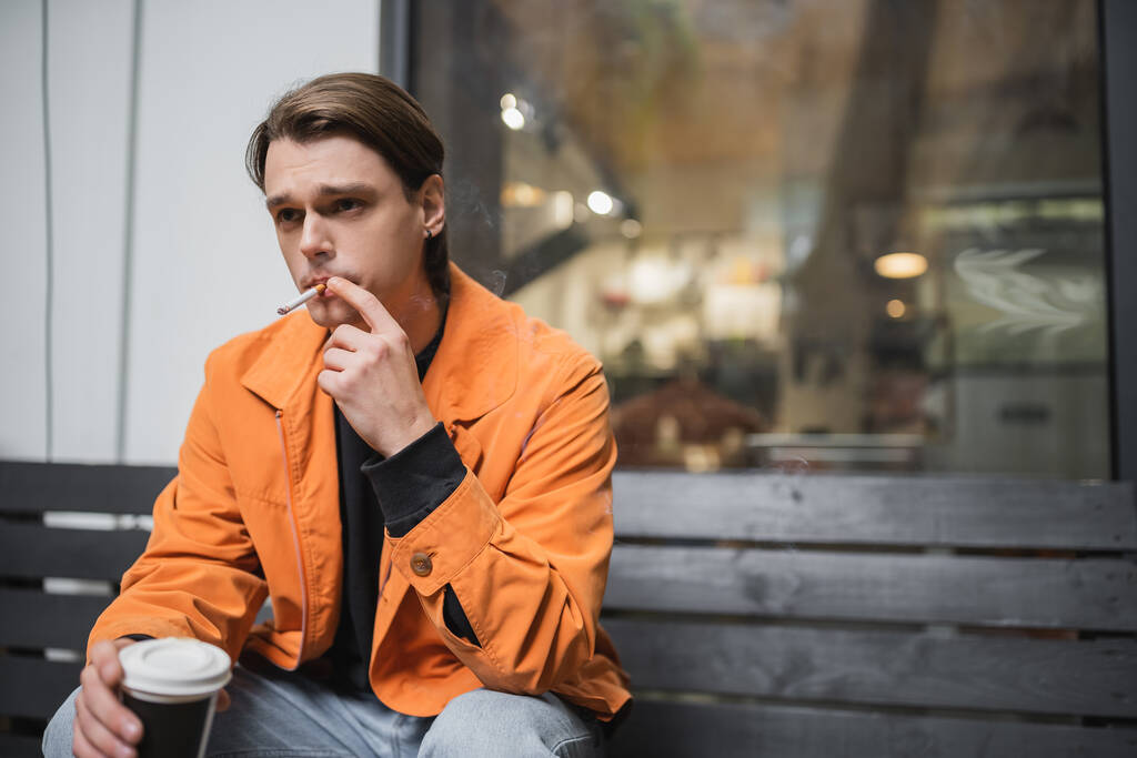 Stylowy człowiek palenia papierosa i trzymając kawę, aby przejść w pobliżu kawiarni  - Zdjęcie, obraz