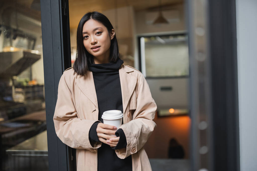 Schöne asiatische Frau im Trenchcoat hält Kaffee in der Nähe der offenen Tür eines Cafés  - Foto, Bild