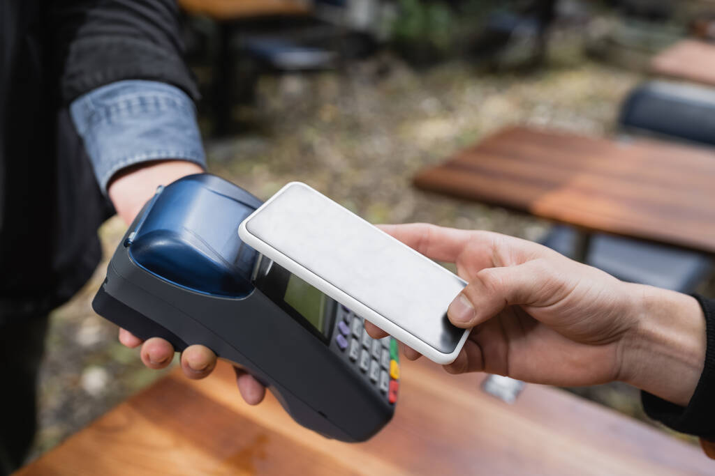 Widok człowieka płacącego komórką w pobliżu kelnerki z czytnikiem kart kredytowych w kawiarni na świeżym powietrzu  - Zdjęcie, obraz