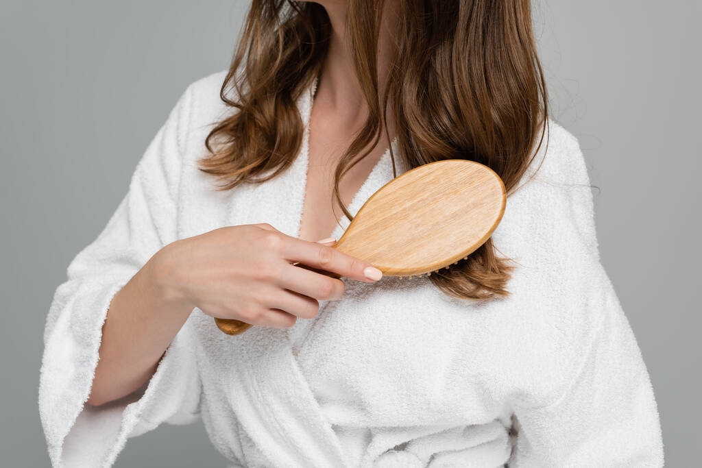 abgeschnittene Ansicht einer jungen Frau im weißen Bademantel, die glänzendes und gesundes Haar isoliert auf grau bürstet - Foto, Bild