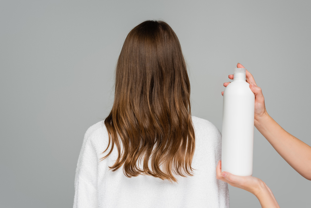 επαγγελματική μαλλιά στυλίστας κρατώντας μπουκάλι με conditioner μαλλιά κοντά σε νεαρή γυναίκα απομονωμένη σε γκρι - Φωτογραφία, εικόνα