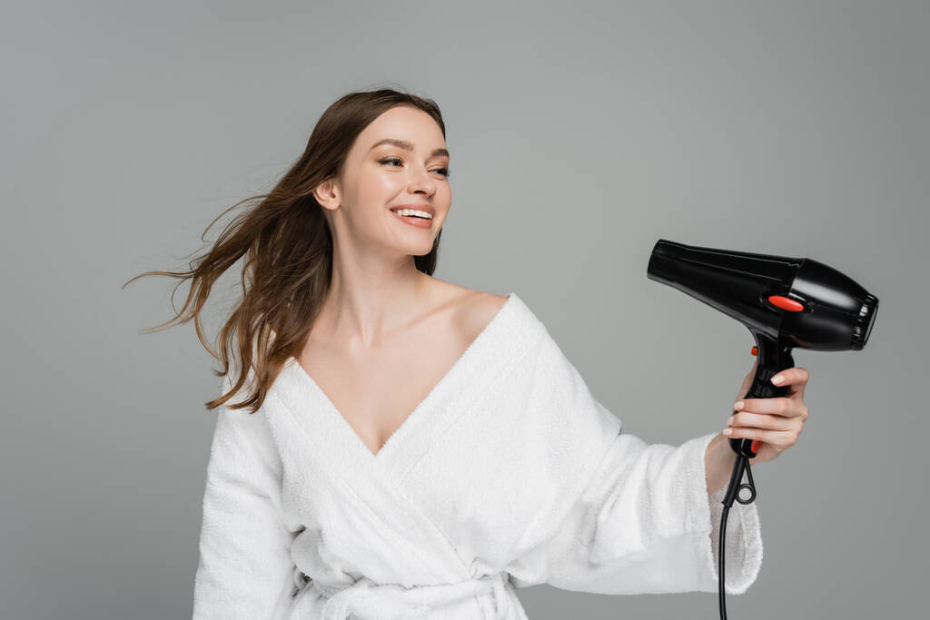 onnellinen nuori nainen kiiltävät hiukset käyttäen hiustenkuivaaja eristetty harmaa  - Valokuva, kuva