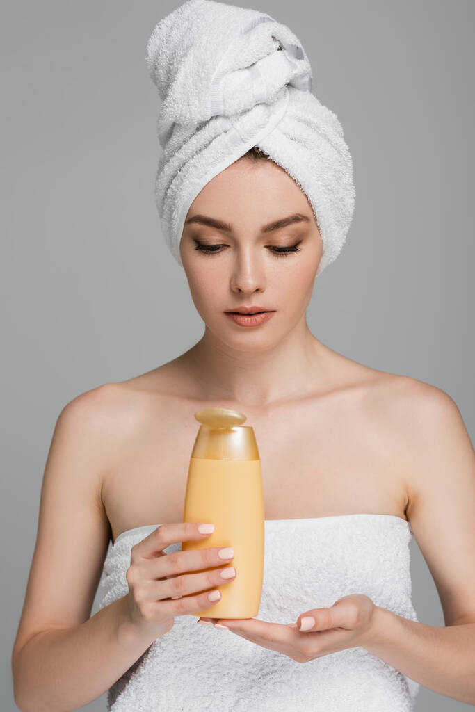 όμορφη γυναίκα με γυμνούς ώμους και πετσέτα στο κεφάλι κρατώντας μπουκάλι με σαμπουάν που απομονώνονται σε γκρι  - Φωτογραφία, εικόνα